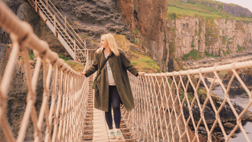 A person walking across Carrick-a-Rede Rope Bridge, with steep cliffs, rocky coastline and the sea visible in the background